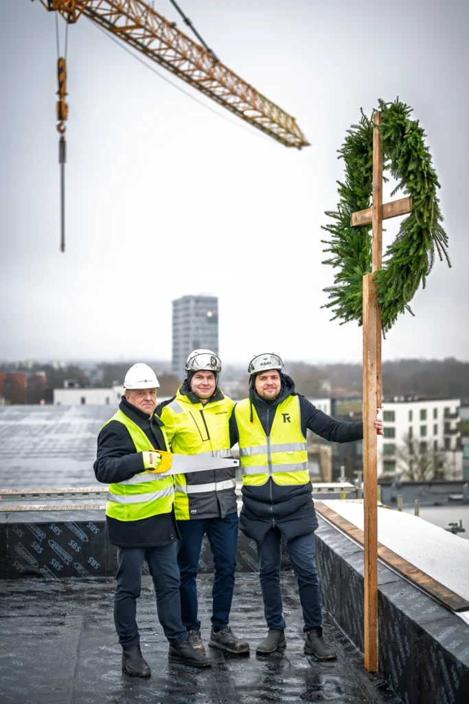Three men in hard hats pose on a rooftop beside a topping-out wreath fixed to a tall wooden pole.
