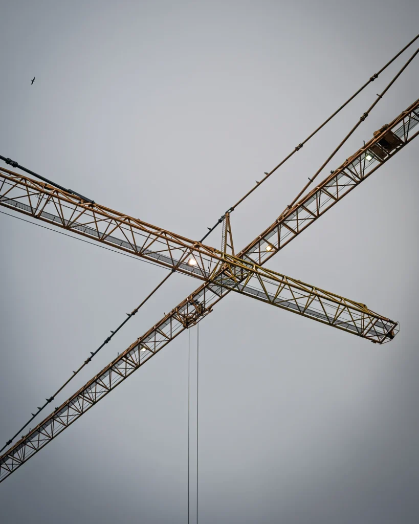 The jibs of two tower cranes cross against a grey sky.