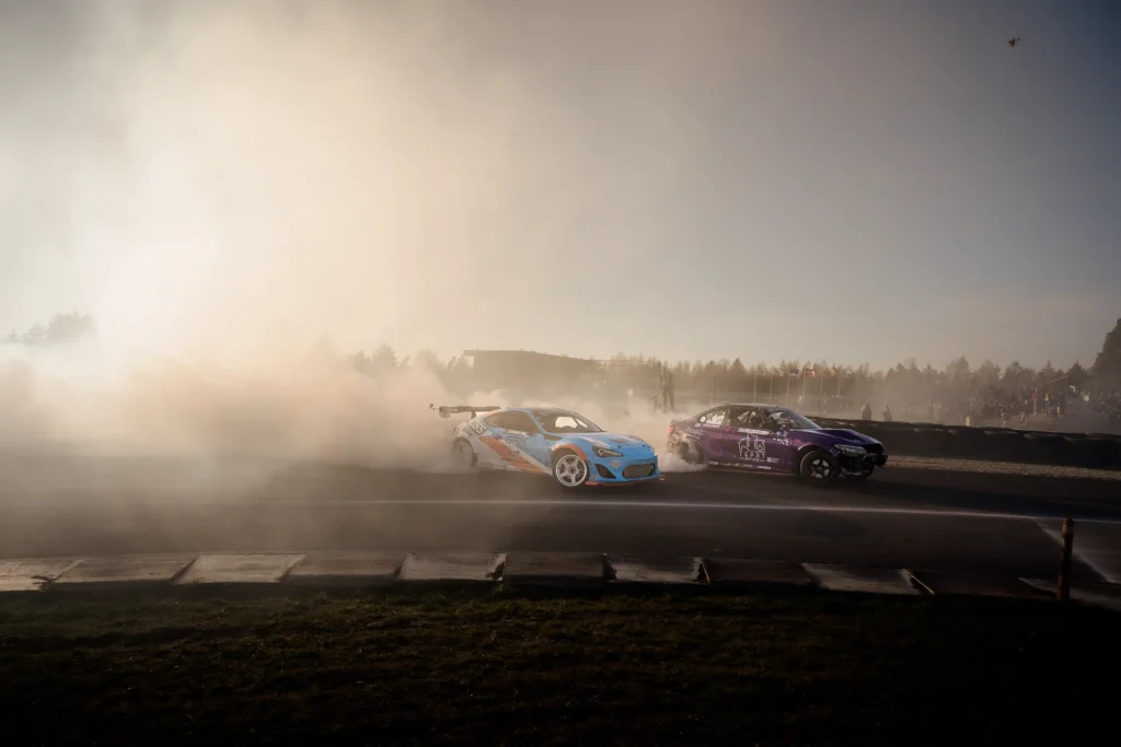 A blue car and a purple car slide sideways through golden, sunlit tyre smoke.