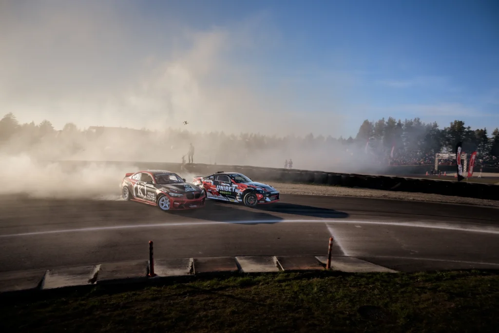 Two red-and-black drift cars run side by side, with smoke and a track barrier behind them.