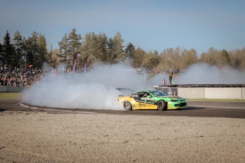 A green-and-yellow drift car corners as smoke partly obscures the spectators behind it.