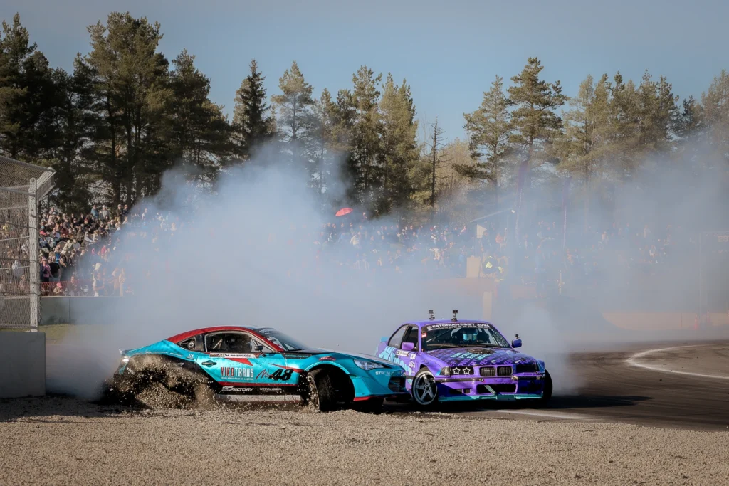 A turquoise drift car throws up gravel at the edge of the track beside a purple car.