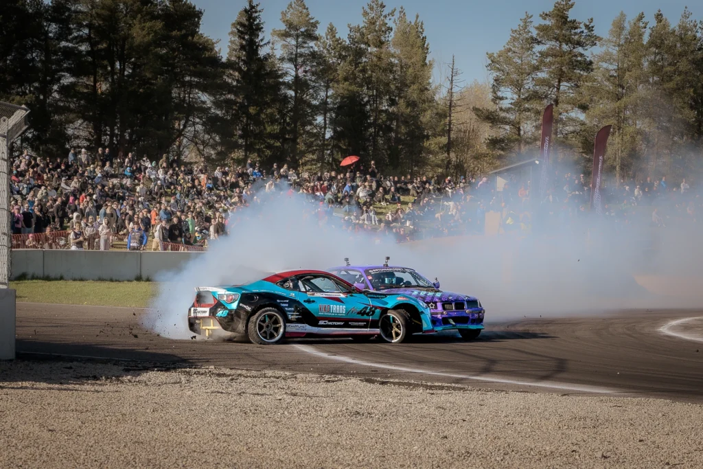 A turquoise car and a purple car drift through a smoky corner in front of the spectators.