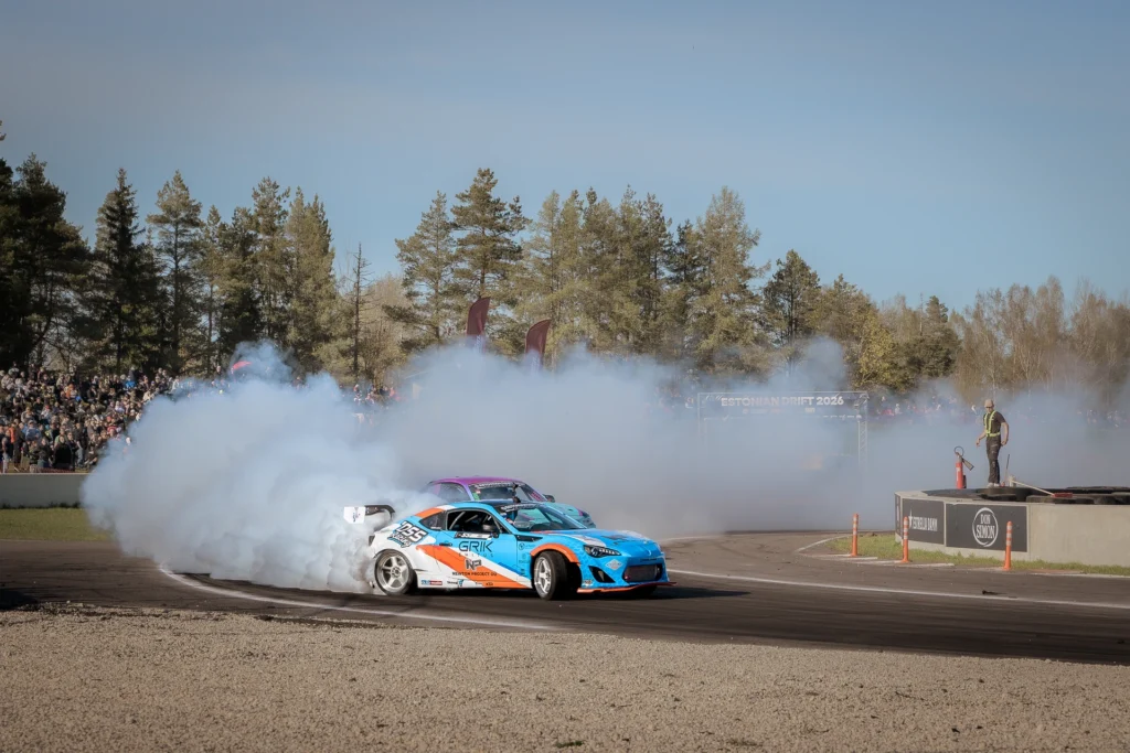 A blue drift car exits a corner with a purple car partly visible behind it.