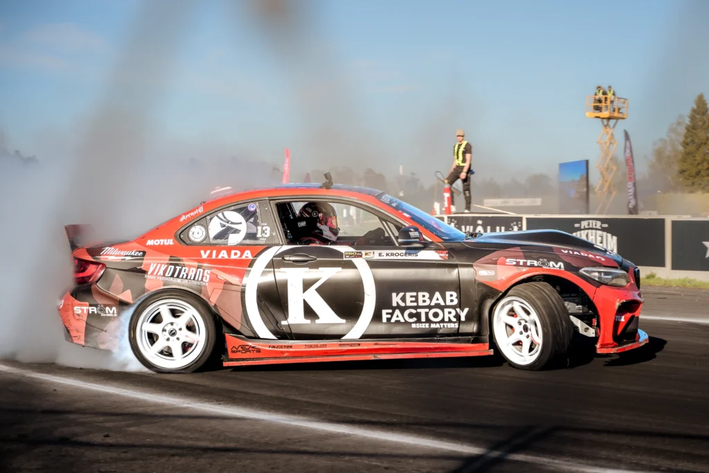 Side view of a red drift car with a large letter K on its door and smoke around the rear wheel.