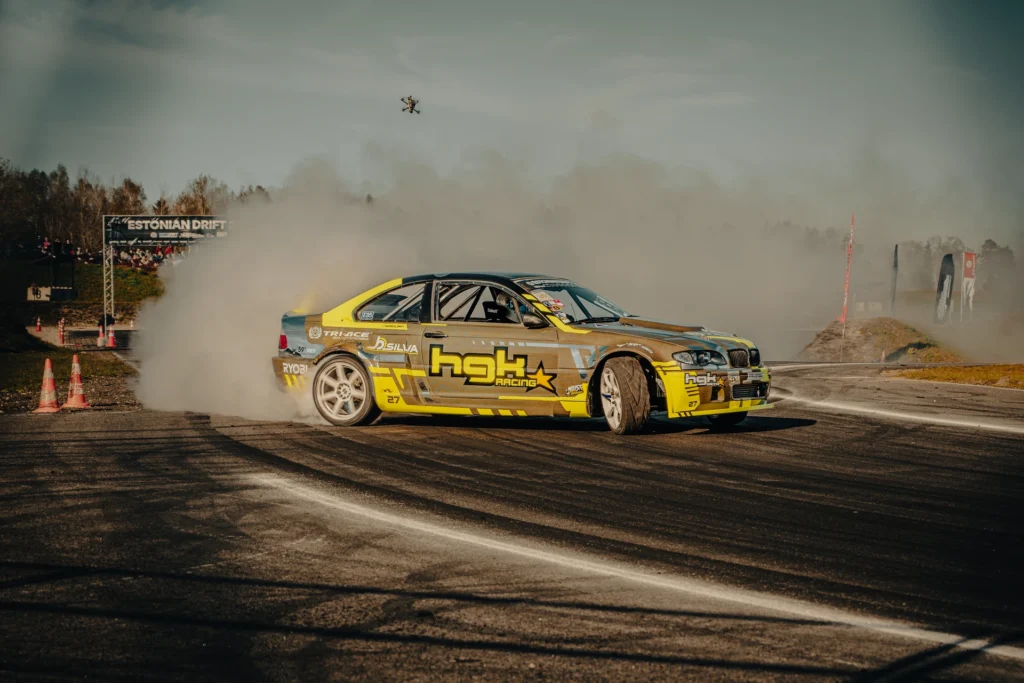 A yellow-and-grey car with white wheels slides along the track, with a drone above the smoke.