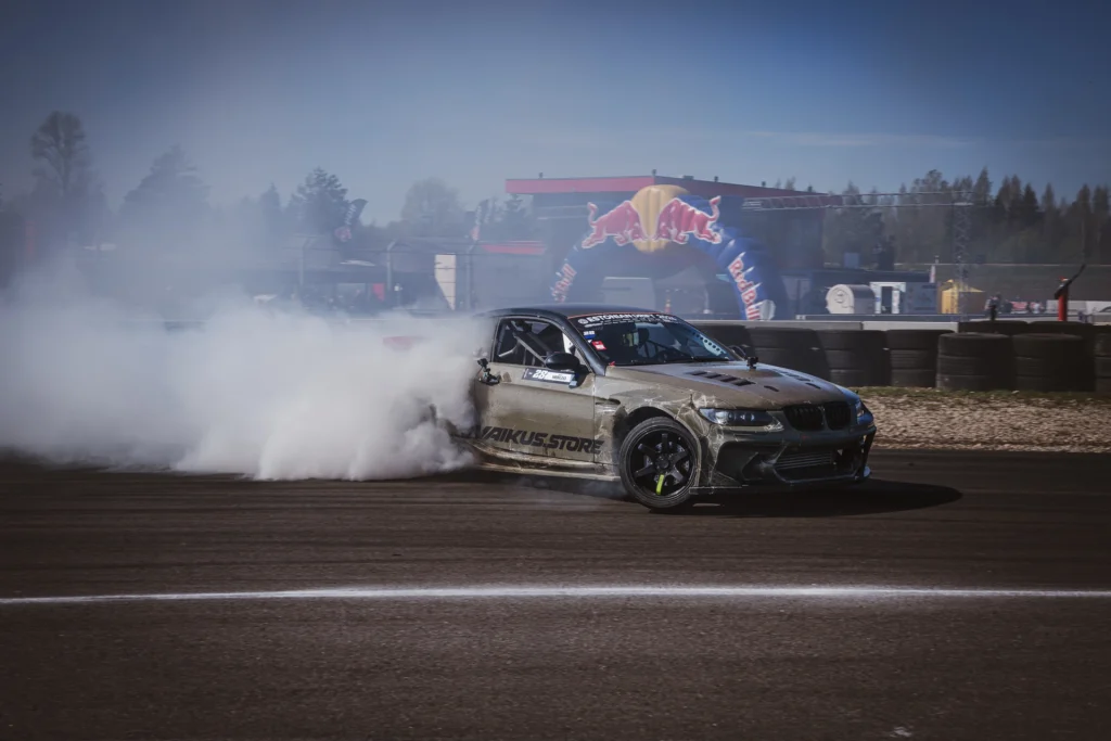 A gold drift car runs ahead of a smoke cloud, with an inflatable event arch in the background.