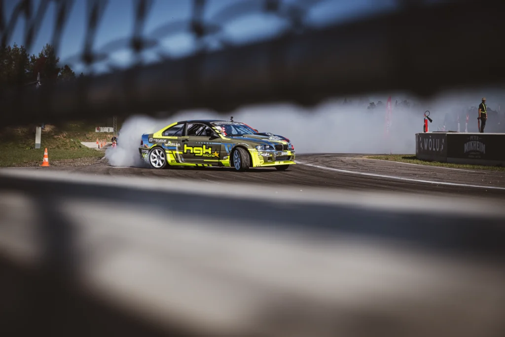 A yellow-and-black drift car on the track, viewed between the horizontal parts of a fence.