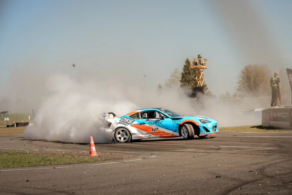 A blue car with a large rear wing slides past a cone in front of a dense smoke cloud.