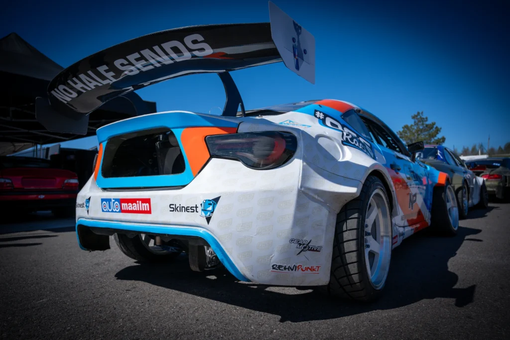 Rear view of a blue-and-orange drift car in the paddock, with a wide rear wing high above the body.