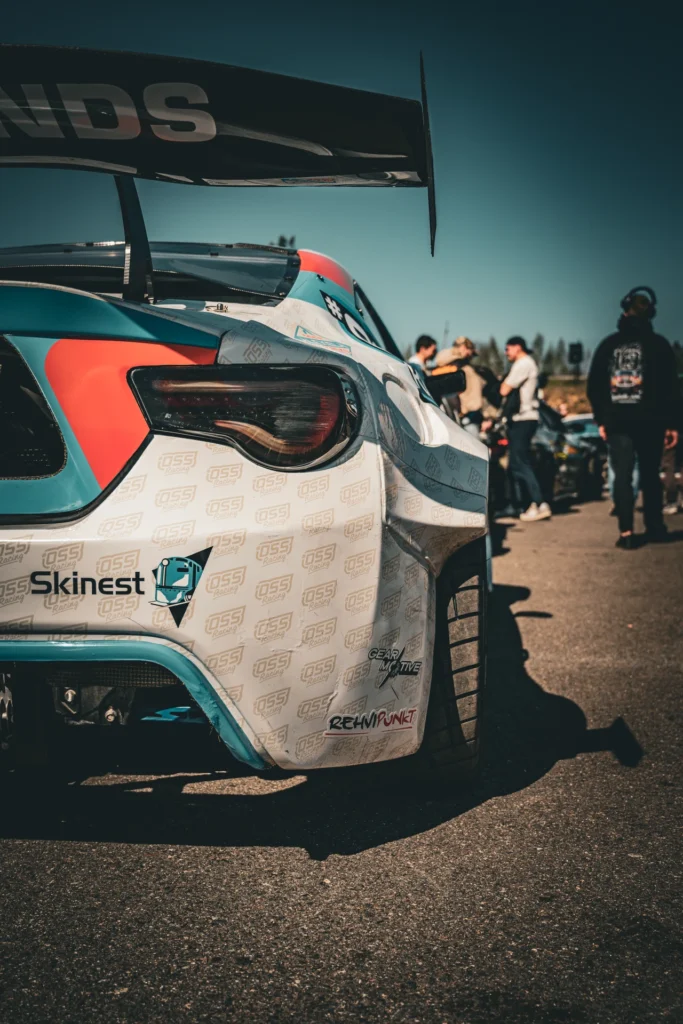 Close view of a white-and-blue drift car's rear corner, with visitors in the paddock behind it.