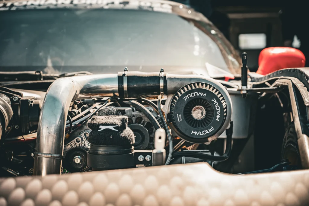 A metal pipe and round black air filter in the engine bay of a car with a red wing mirror.