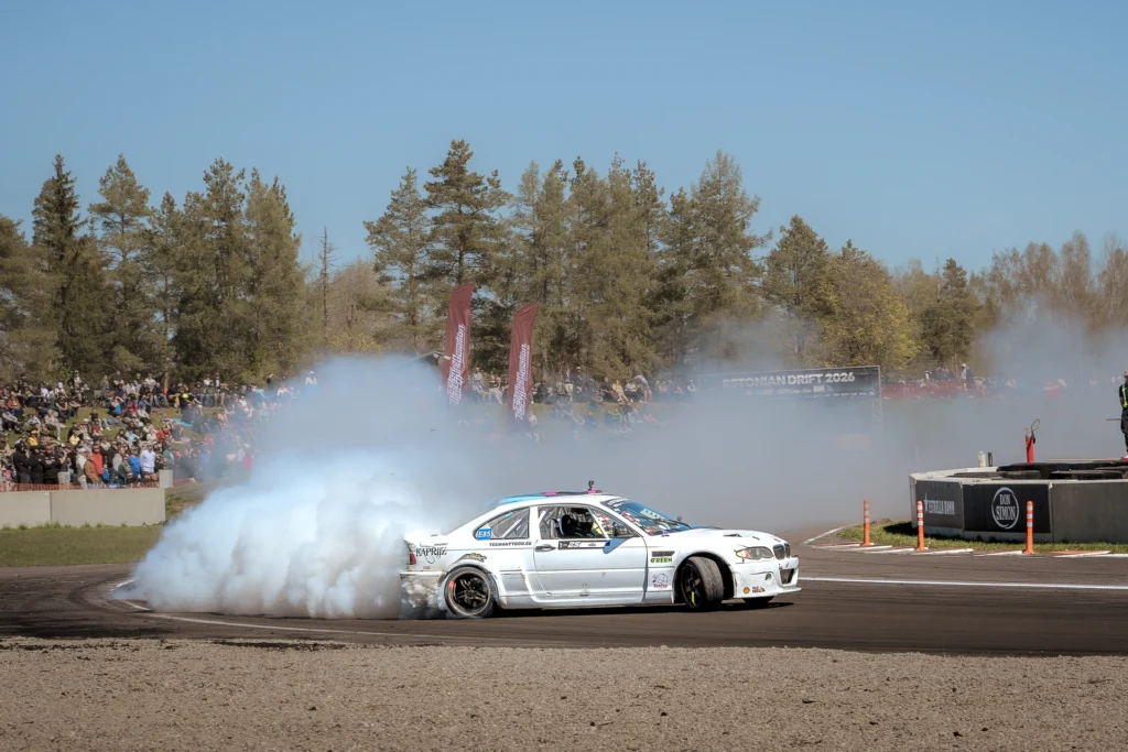 A white drift car slides through a corner with dense tyre smoke behind its rear wheels.