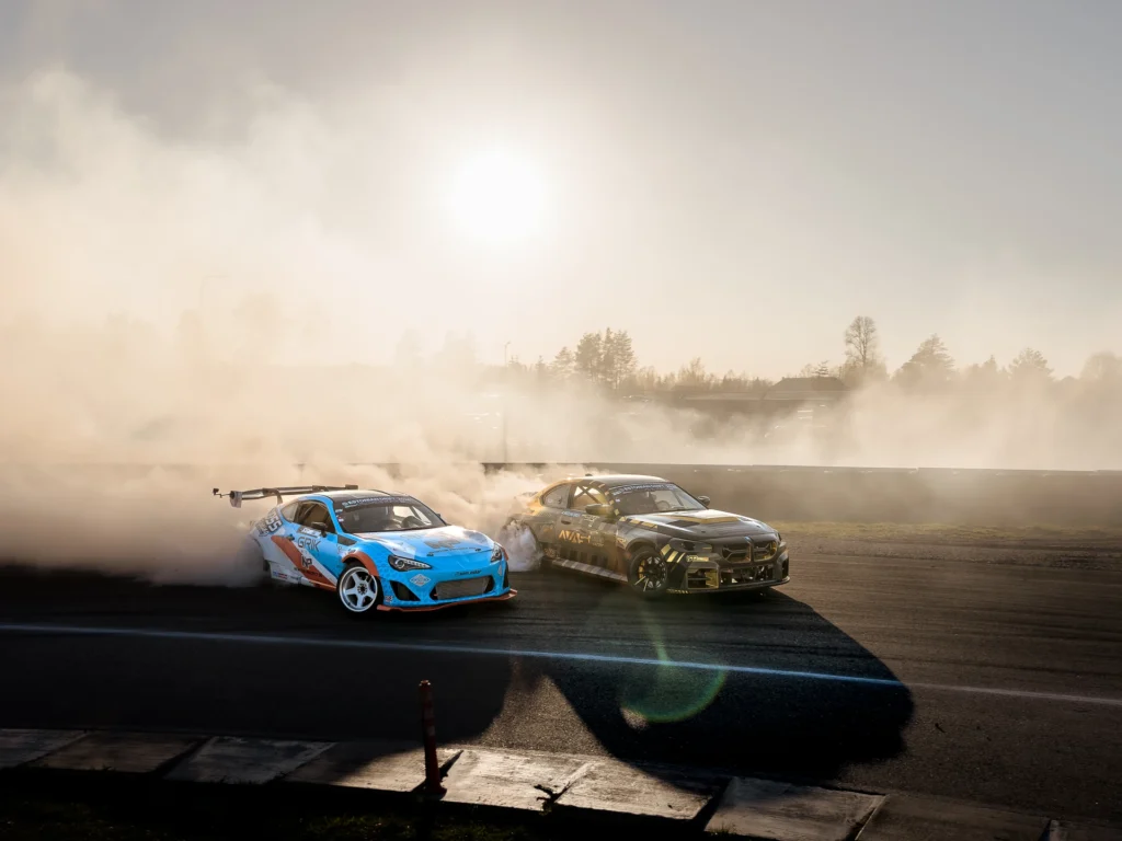 Two cars drifting side by side cast long shadows in sunlight filtering through the smoke.
