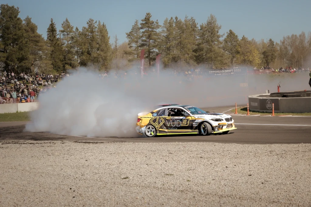 A yellow-and-black drift car with a white front end runs along the outside of a smoky bend.