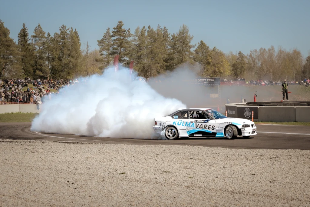 A white drift car with light-blue lettering trails a tall cloud of tyre smoke.