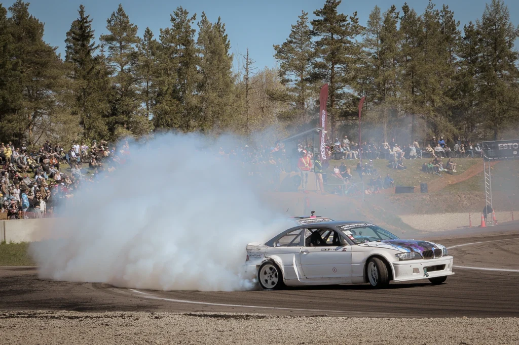 A silver drift car corners with gravel in the foreground and spectators behind the smoke.