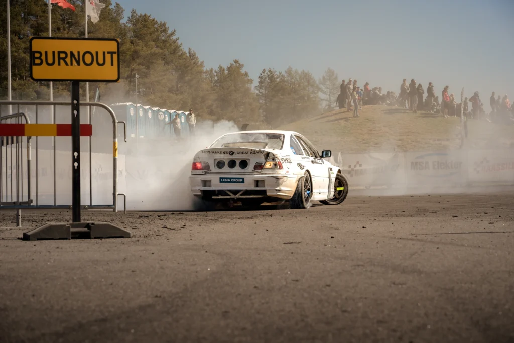 Rear view of a white drift car in tyre smoke beside a yellow sign reading BURNOUT.
