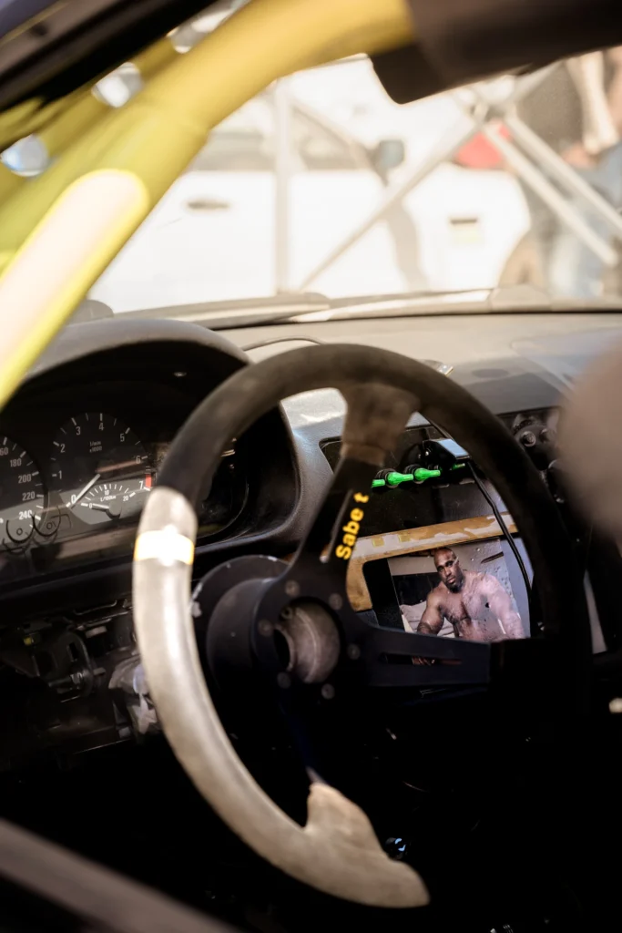 A racing steering wheel with worn covering and a small photograph attached, beside a yellow roll cage.