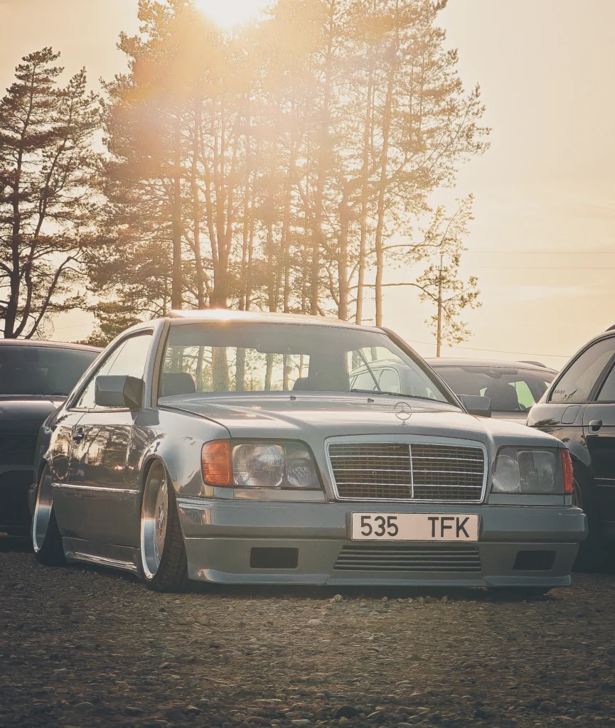 A lowered silver car parked on grass, with sunlight shining through pine trees behind it.