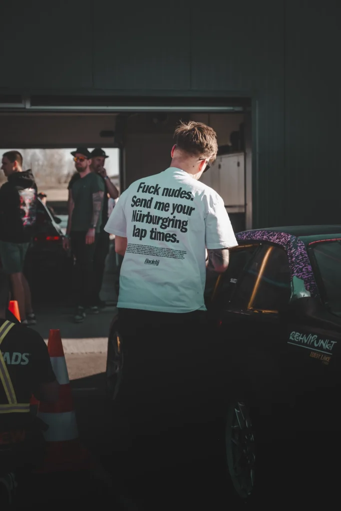 A person wearing a light-coloured T-shirt with lettering stands beside a racing car in the paddock.