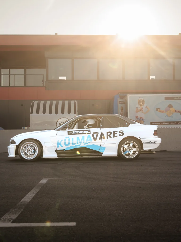 Side view of a white drift car in front of a building, with low sunlight above the roof.