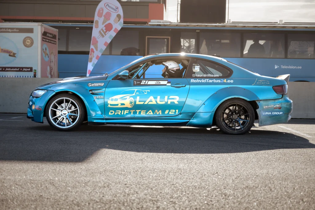 Side view of a turquoise drift car in the paddock, with a light front wheel and black rear wheel.