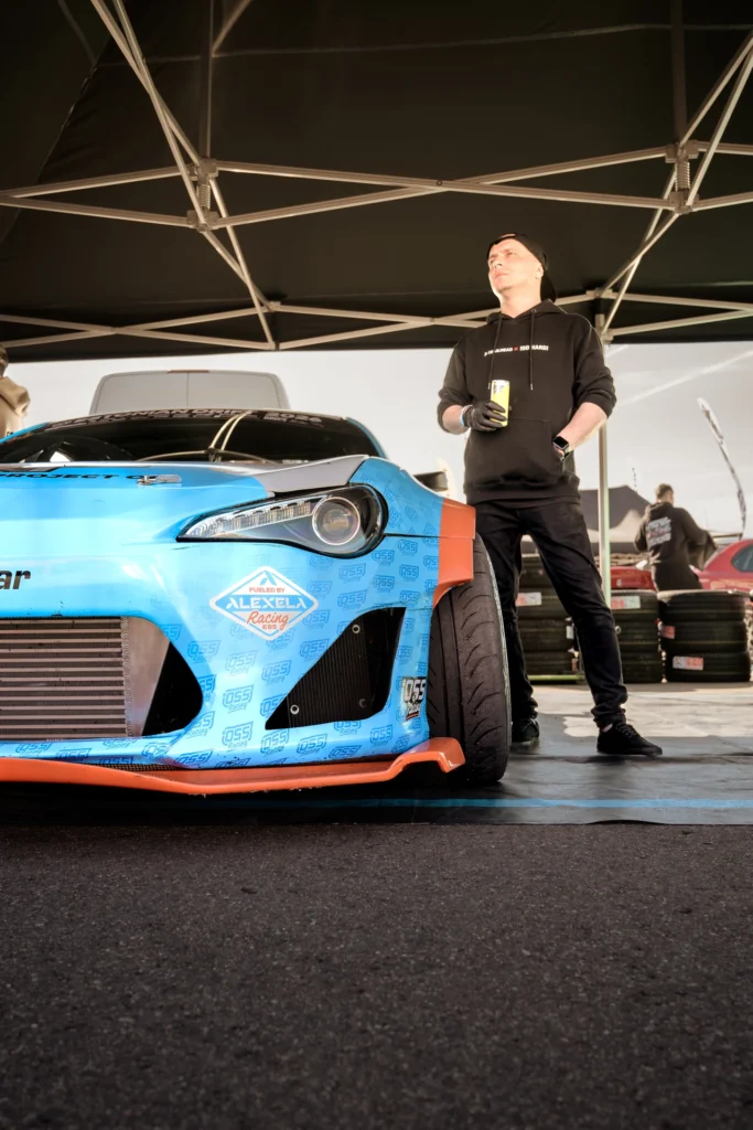 The front corner of a blue drift car beside a person holding a drink under a paddock tent.