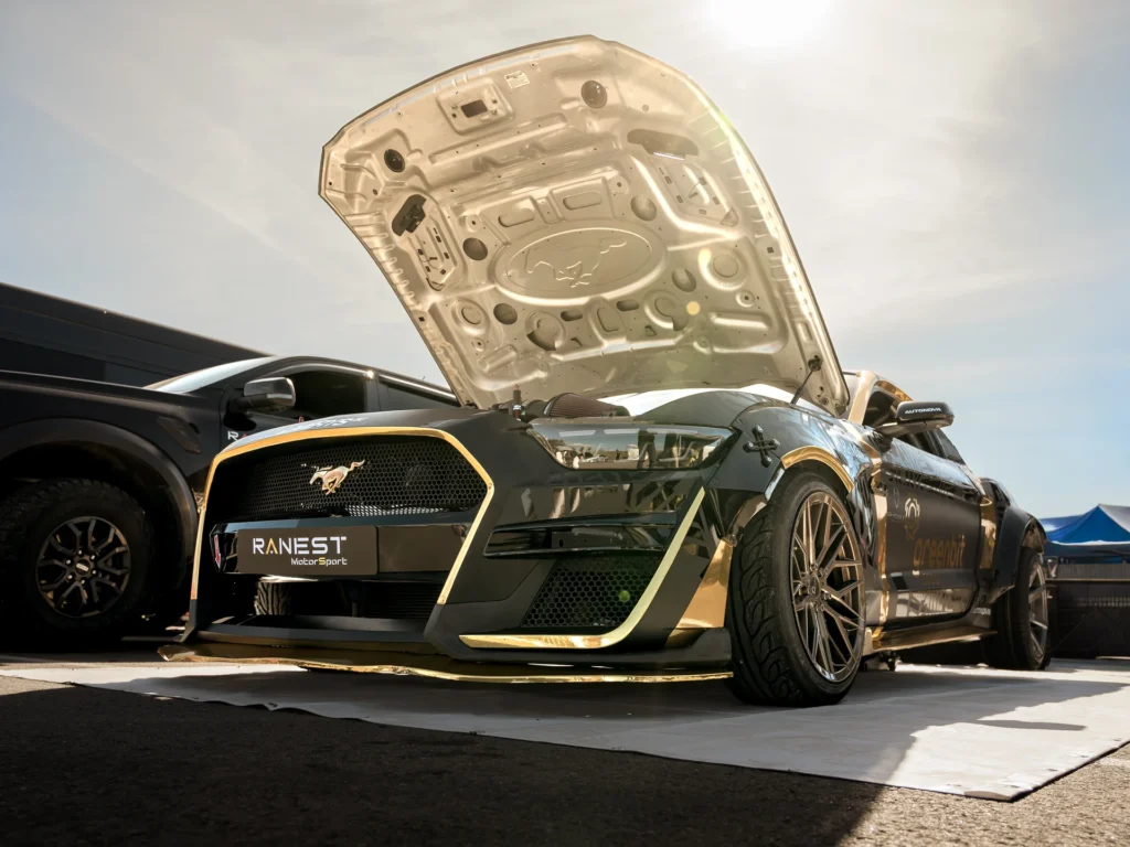 Low front view of a black-and-gold racing car with its bonnet open and a light mat beneath it.