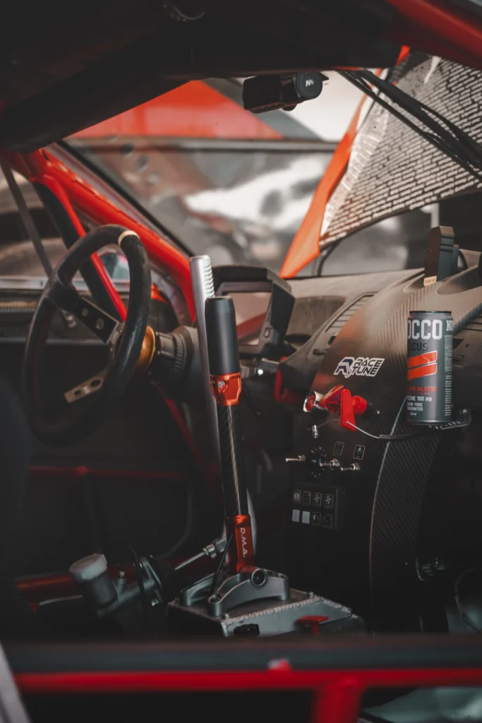 A racing car cockpit with a red roll cage and tall control levers beside the steering wheel.
