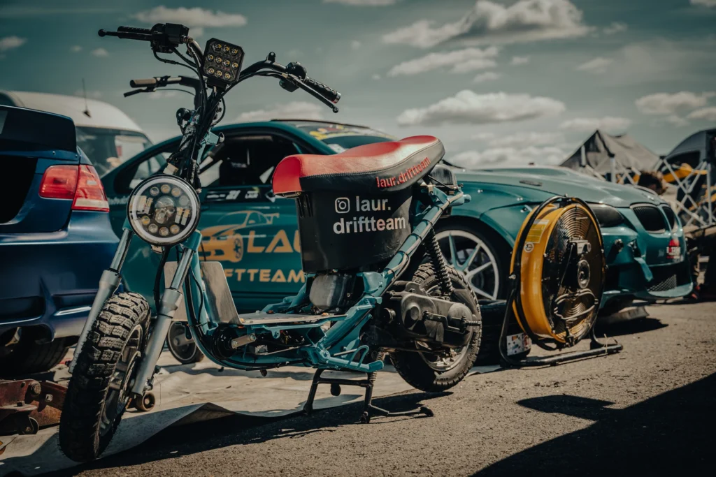 A small moped with a red seat between drift cars, beside a yellow fan.