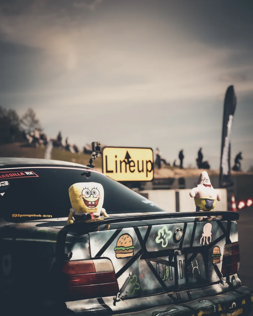 Two toy figures sit on a drift car's large rear wing, with a Lineup sign behind them.