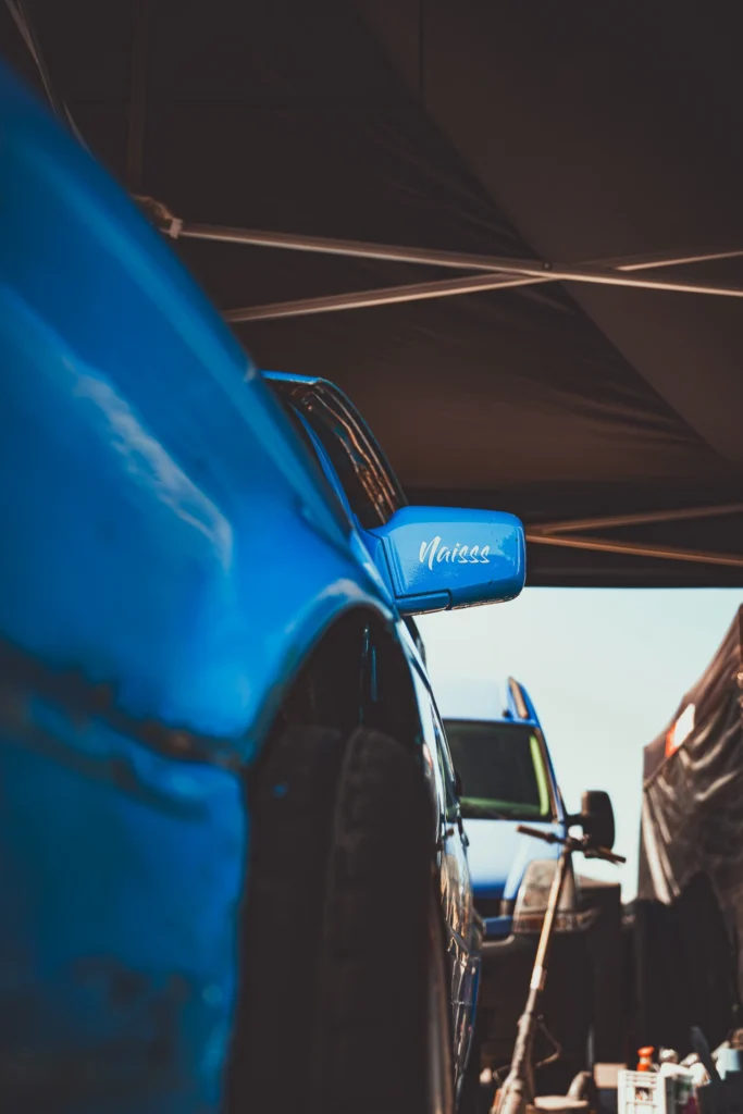 Low-angle view of a blue racing car's wide wheel arch and wing mirror beneath a paddock tent.