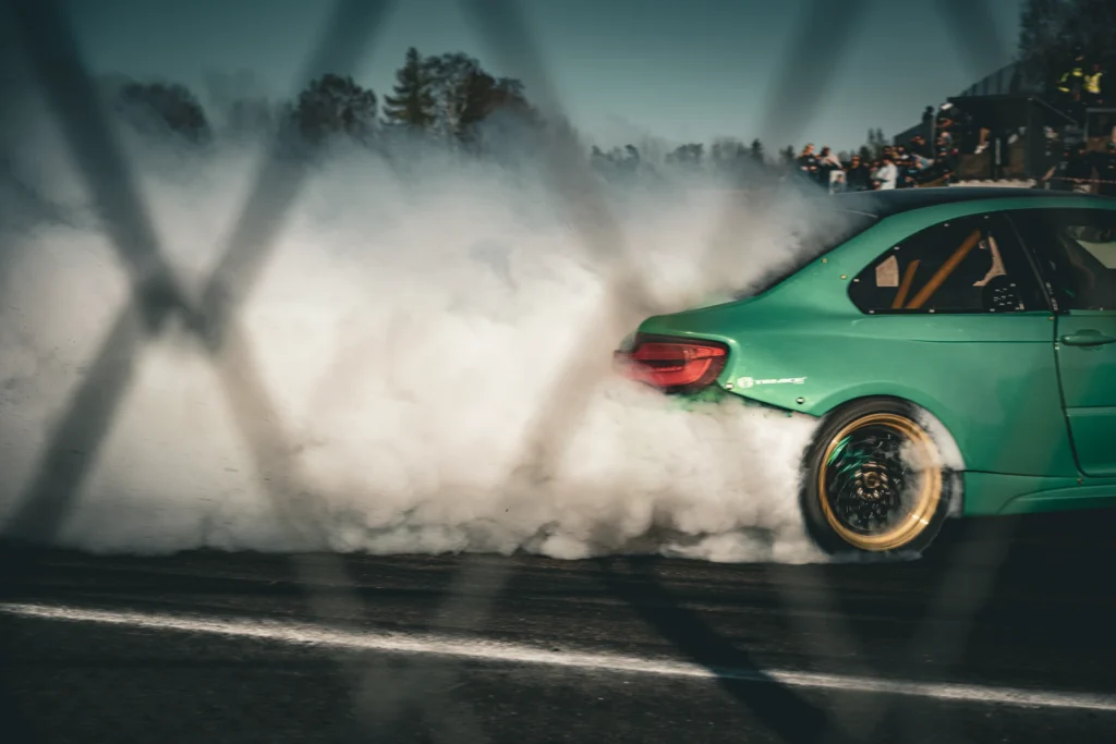 Close view through a fence of a green drift car's rear quarter and smoking rear tyre.