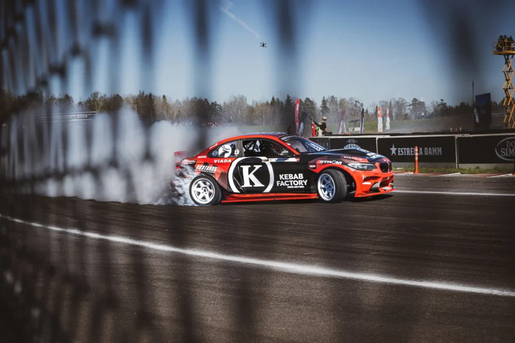 A red drift car seen through a fence, its mesh blurred along the left edge of the frame.