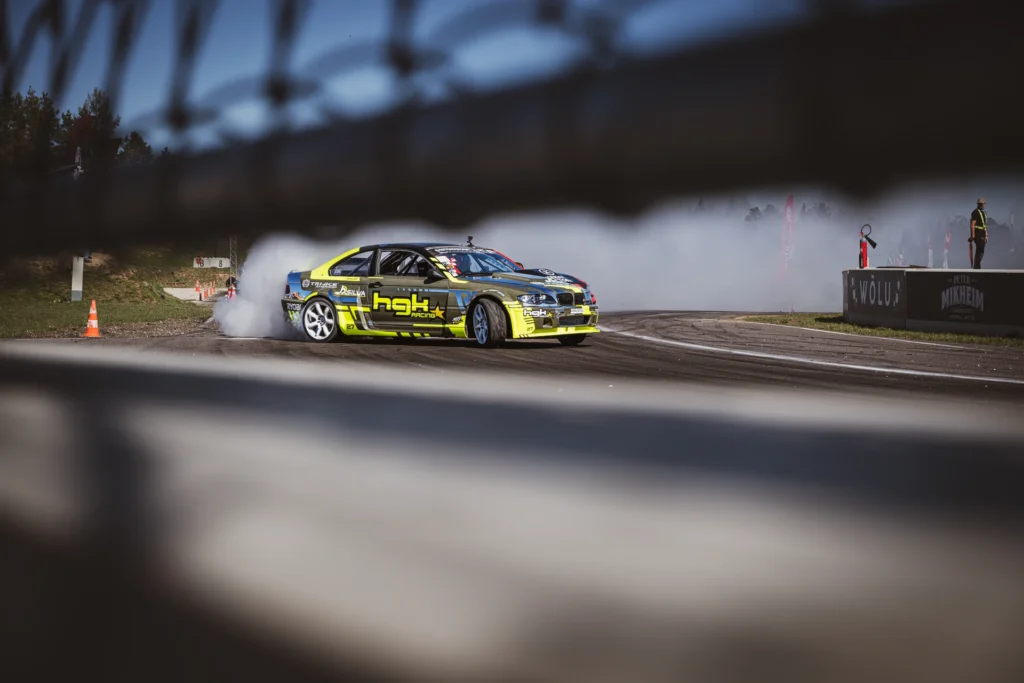 A yellow-and-black car slides through a corner, framed above and below by a blurred fence.