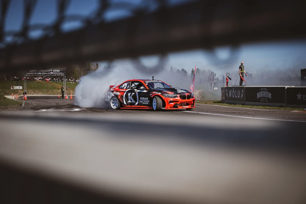 A red drift car in white tyre smoke, photographed through a gap in a fence.