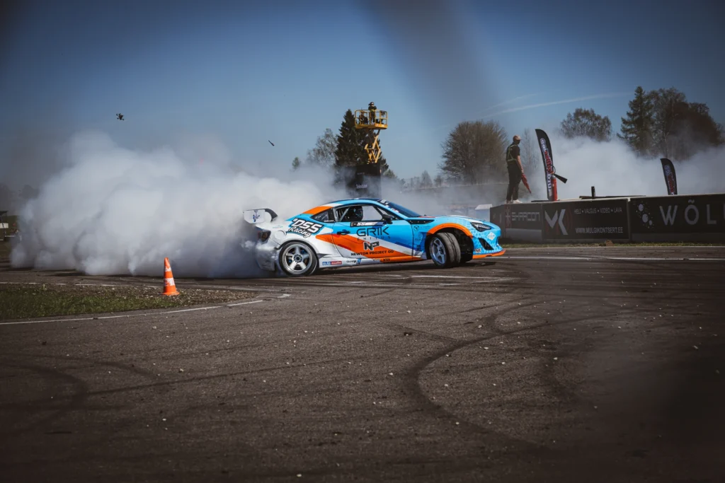 A blue-and-orange drift car beside an orange cone, with an aerial lift and tyre smoke behind it.