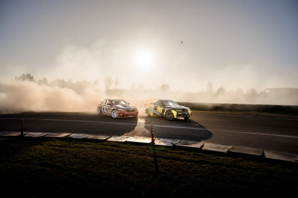 A red car and a yellow-and-black car tandem drift across the sunlit circuit.