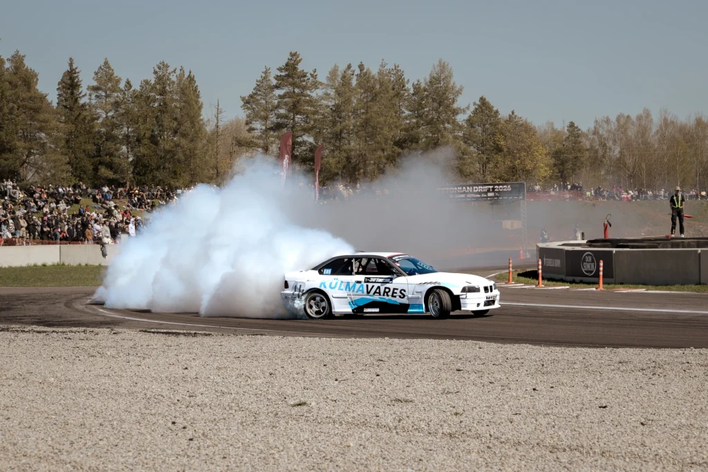 A wide track view of a white drift car, with smoke and spectators seated in front of pine trees.