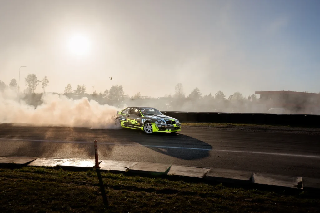 A green-and-white drift car trails a long plume of smoke in the low sunlight.