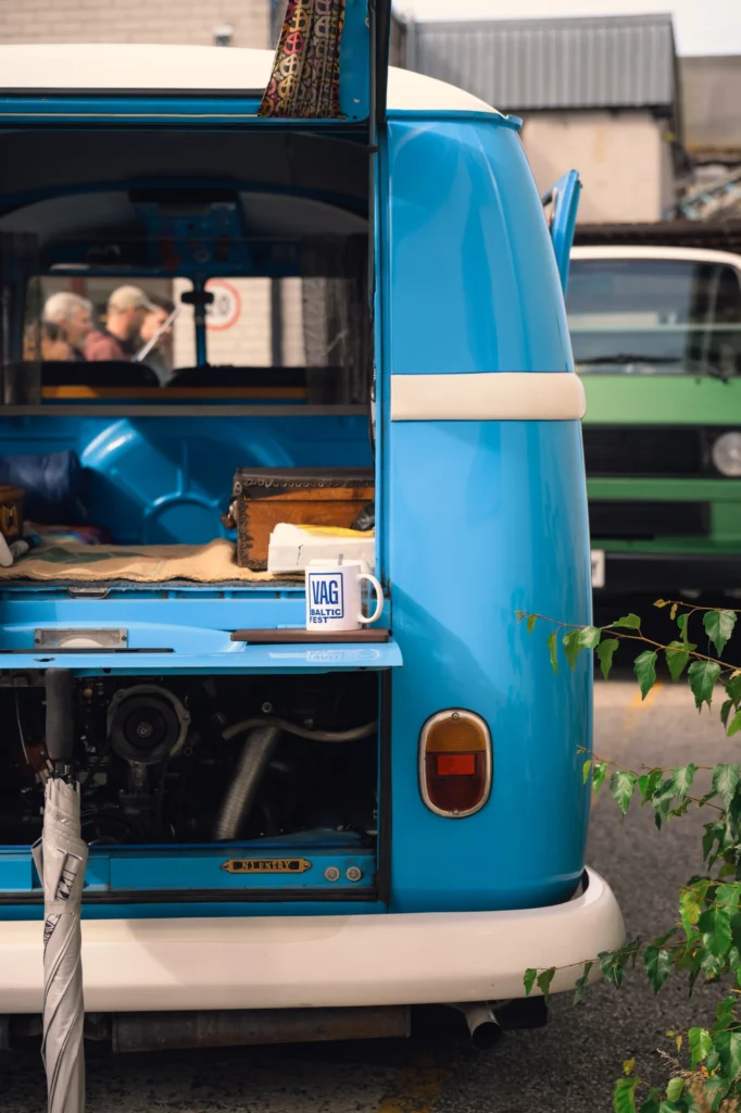 Open rear hatch of a blue and white campervan, with a mug on the ledge and the engine compartment below.