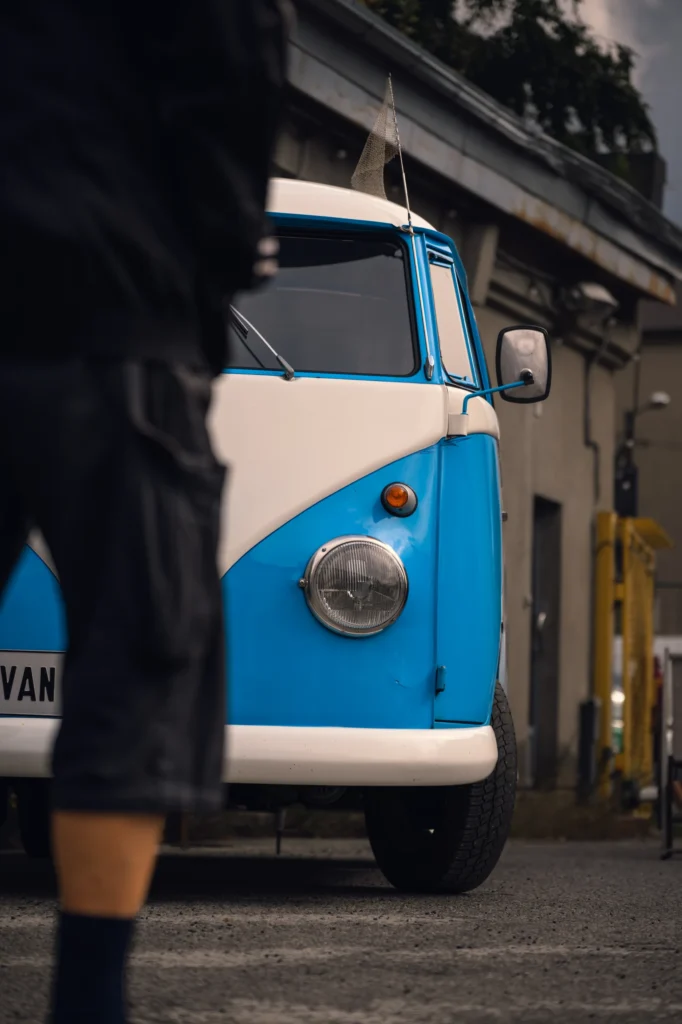 The front of a blue and white Volkswagen outside a factory building, partly obscured by a passer-by.