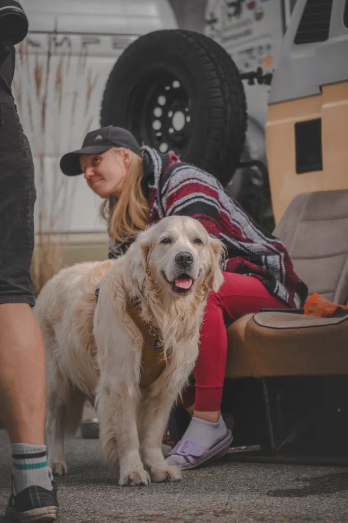 A pale long-haired dog stands in front of a woman in a striped poncho seated beside a campervan.