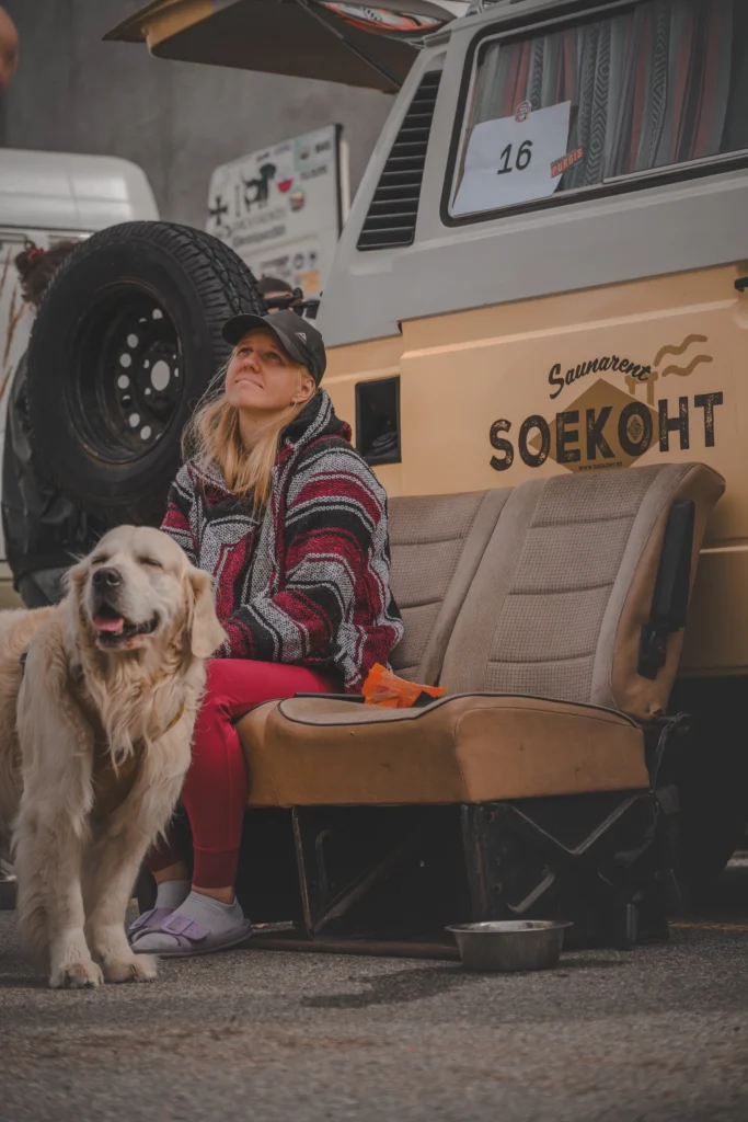 A woman in a striped poncho sits on a car seat beside a dog, with a beige campervan and spare wheel behind them.