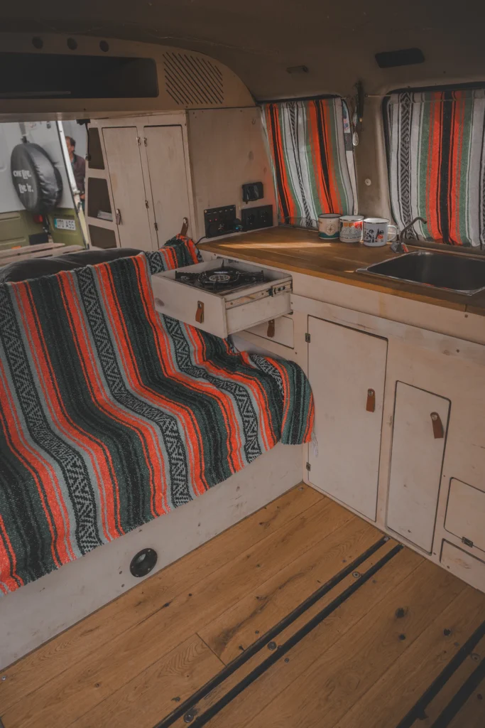 A campervan kitchenette with light-coloured cabinets, a sink and a stove beside a seat covered in red and black stripes.