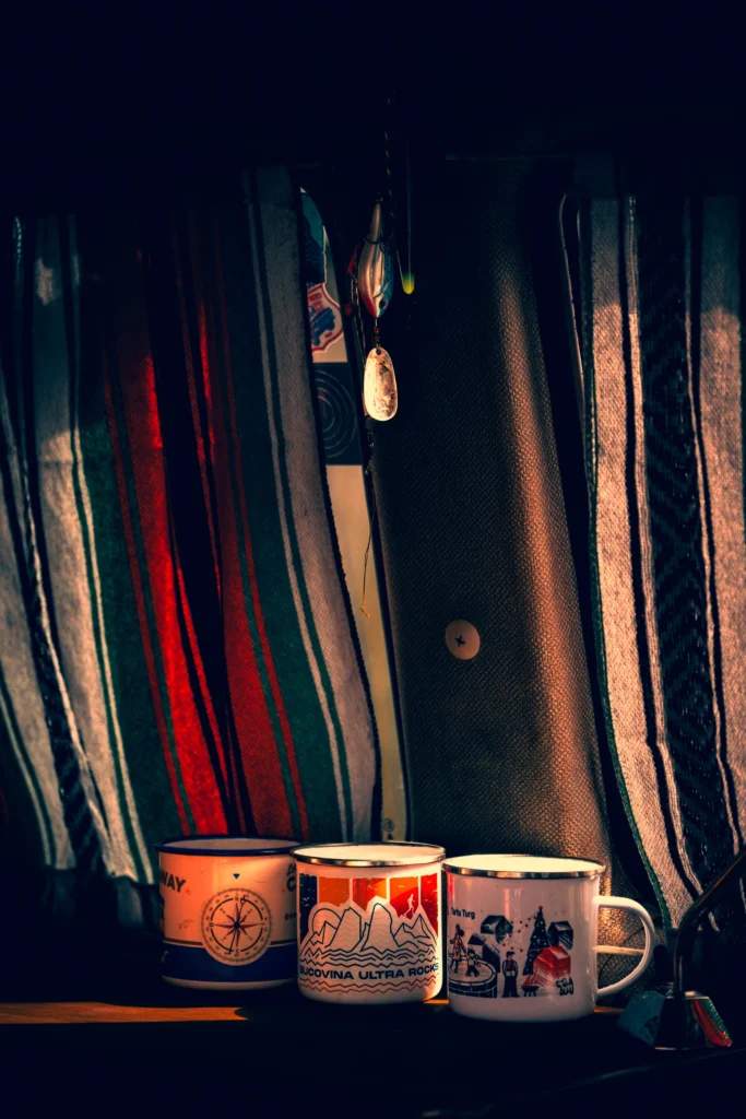 Three illustrated enamel mugs in front of striped curtains, lit by a shaft of light from the side.