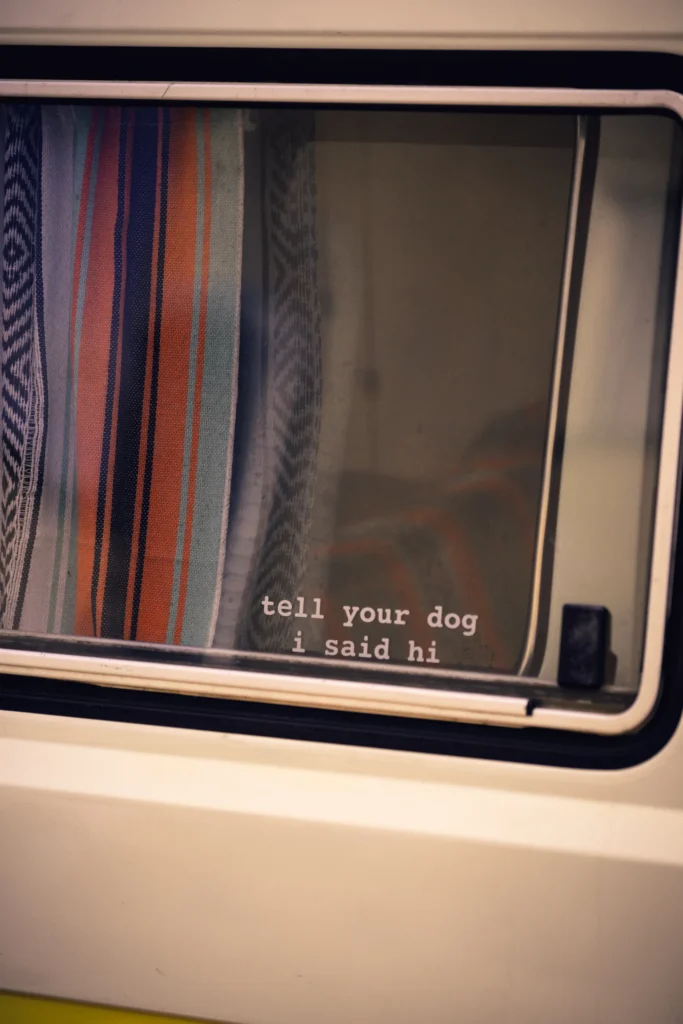 A campervan window with a striped curtain and a sticker reading “tell your dog i said hi”.