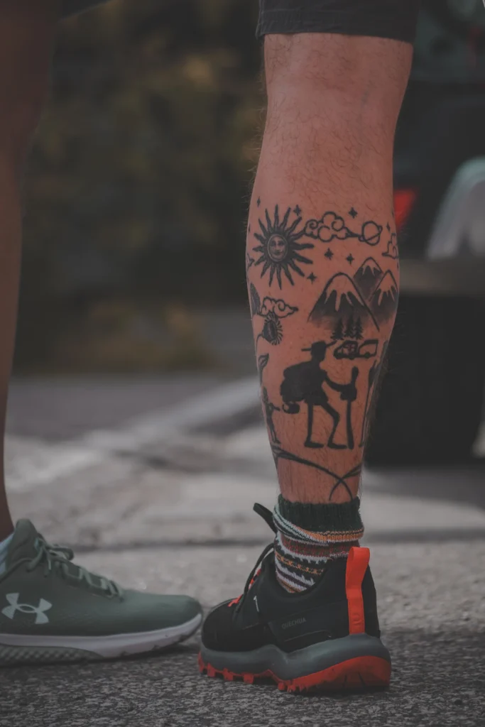 Close-up of a calf tattoo showing a hiker, mountains, the sun and clouds, above dark outdoor shoes.