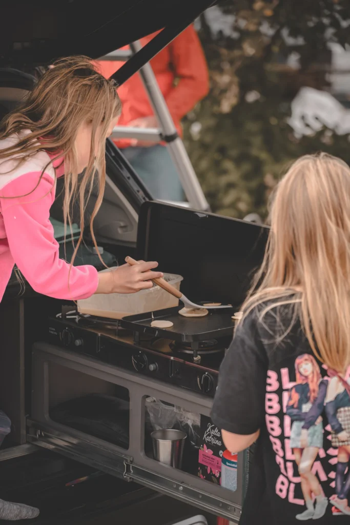 Two children at a car's open tailgate, one spooning batter onto the griddle of a pull-out kitchen unit.